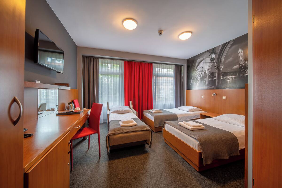 Habitación de hotel con dos camas individuales, un escritorio con silla, televisor y un ventanal con cortinas.