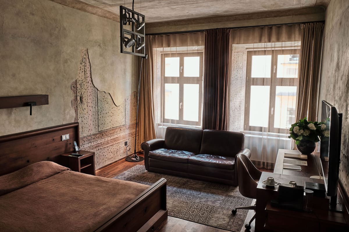 A bedroom with a bed, a leather sofa, a desk with a computer, and a large window with curtains.