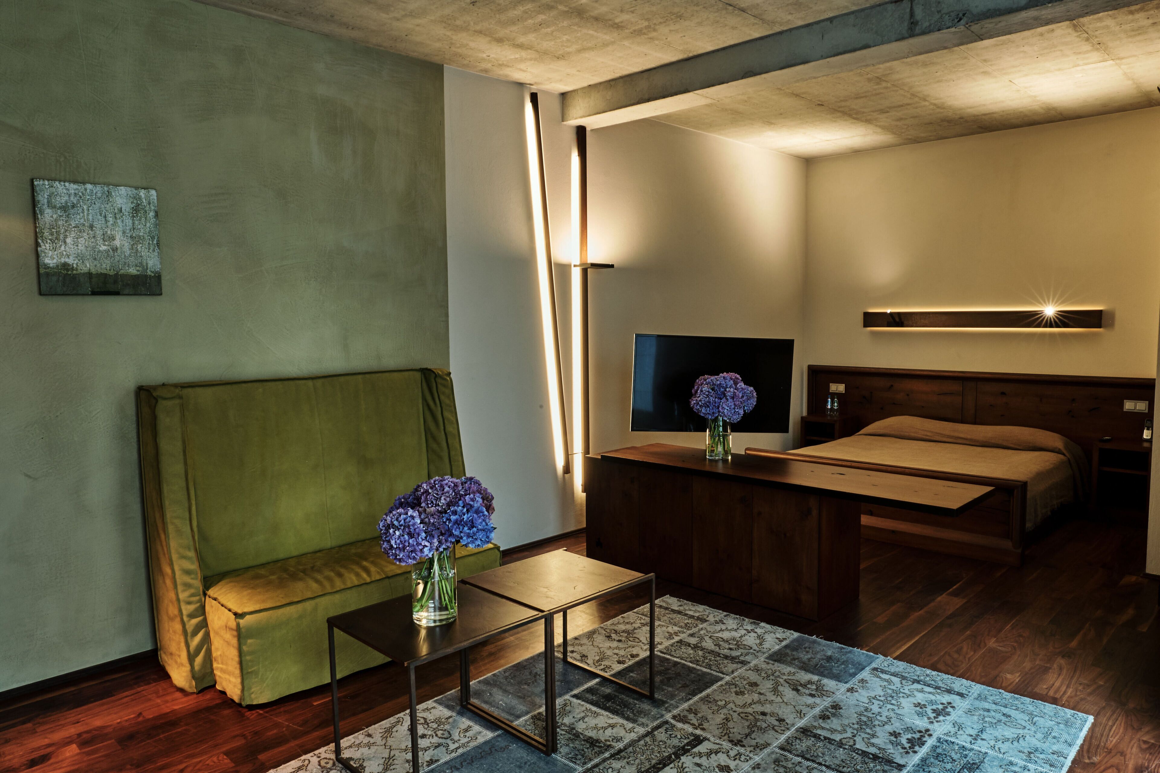 A modern hotel room with a green sofa, a small table with a vase of flowers, a bed, and a TV.