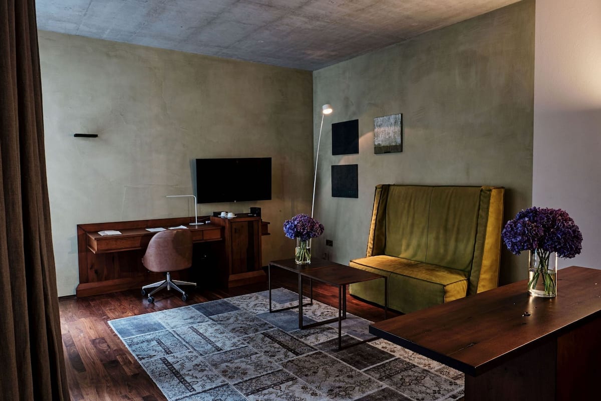 A modern living room with a wooden desk, a green sofa, a flat-screen TV mounted on the wall, and a floor lamp.