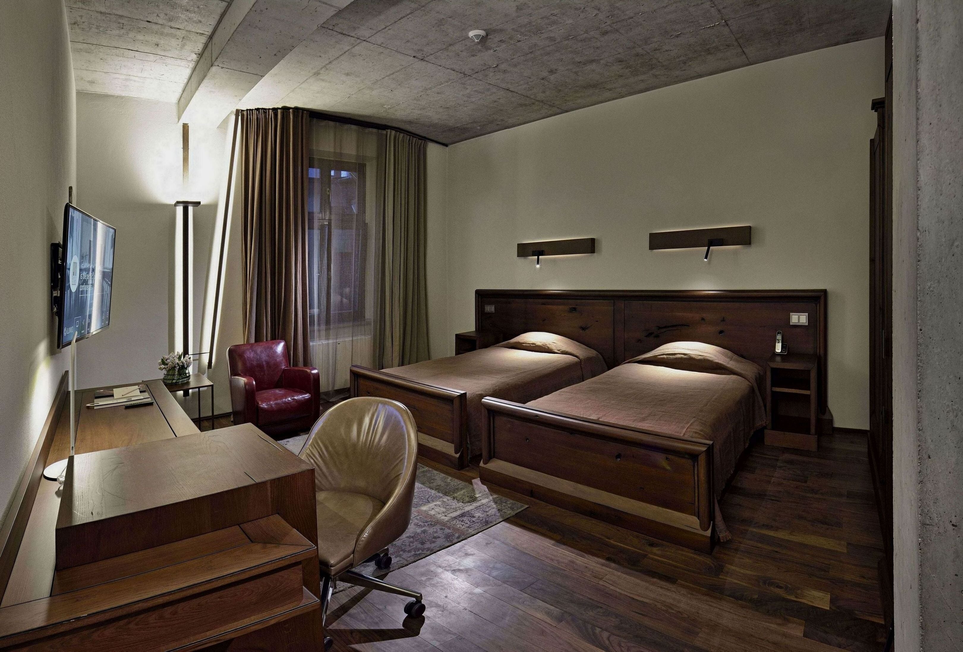 Une chambre d’hôtel avec un lit en bois, un bureau avec une chaise, une télévision et une grande fenêtre avec des rideaux.