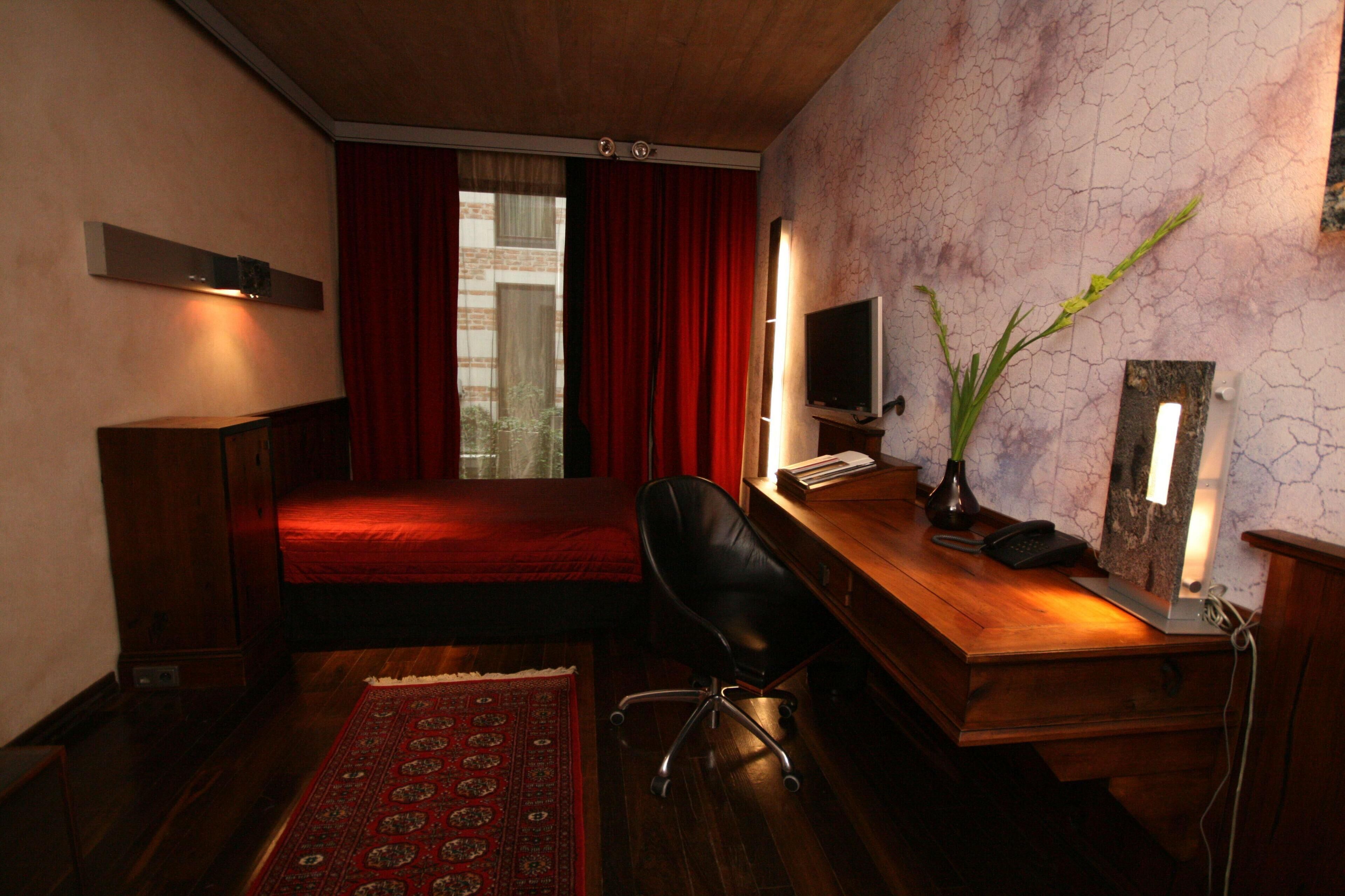 Habitación pequeña con cama, escritorio con computadora, silla, televisor y cortina roja.