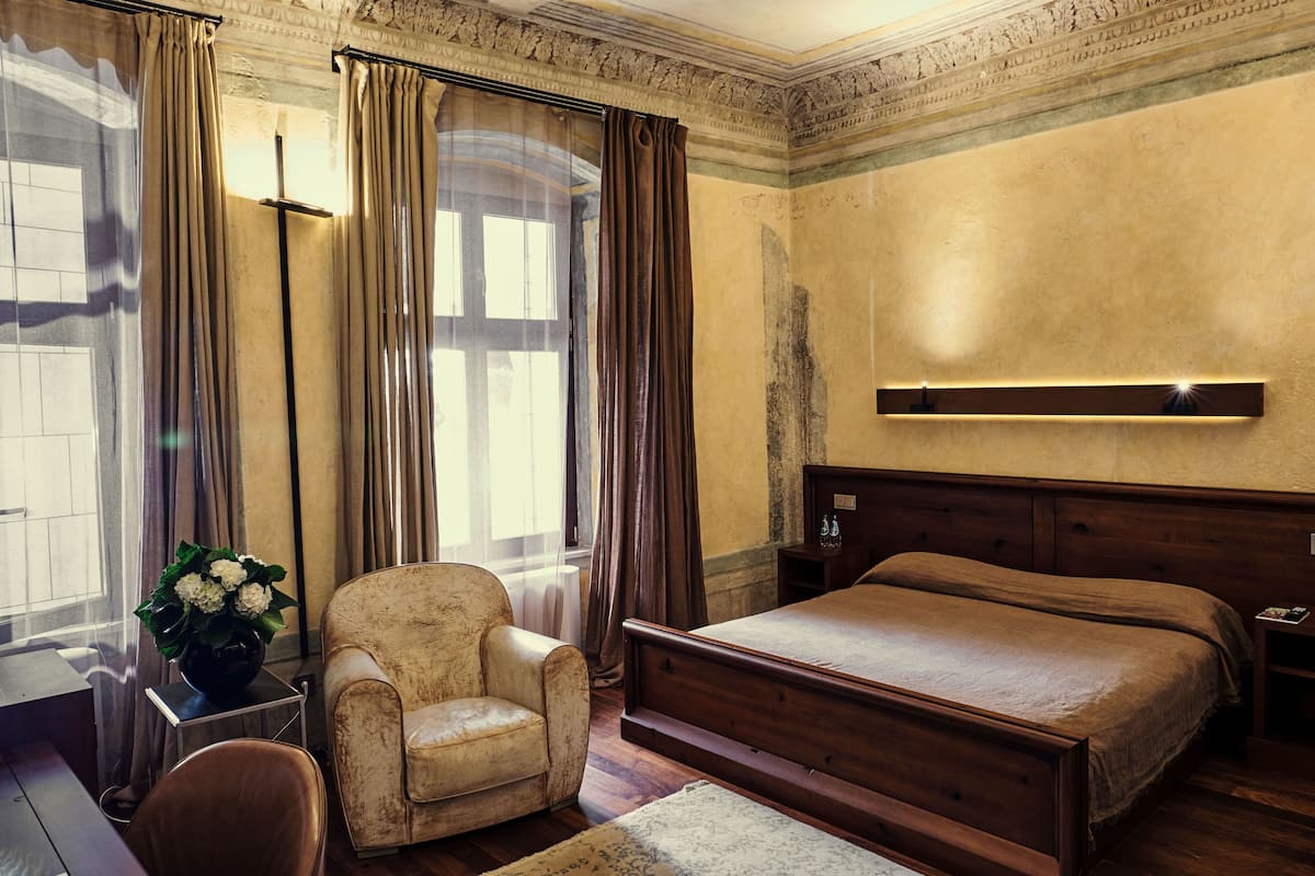 A bedroom with a wooden bed, a chair, a desk with a vase of flowers, and a large window with curtains.