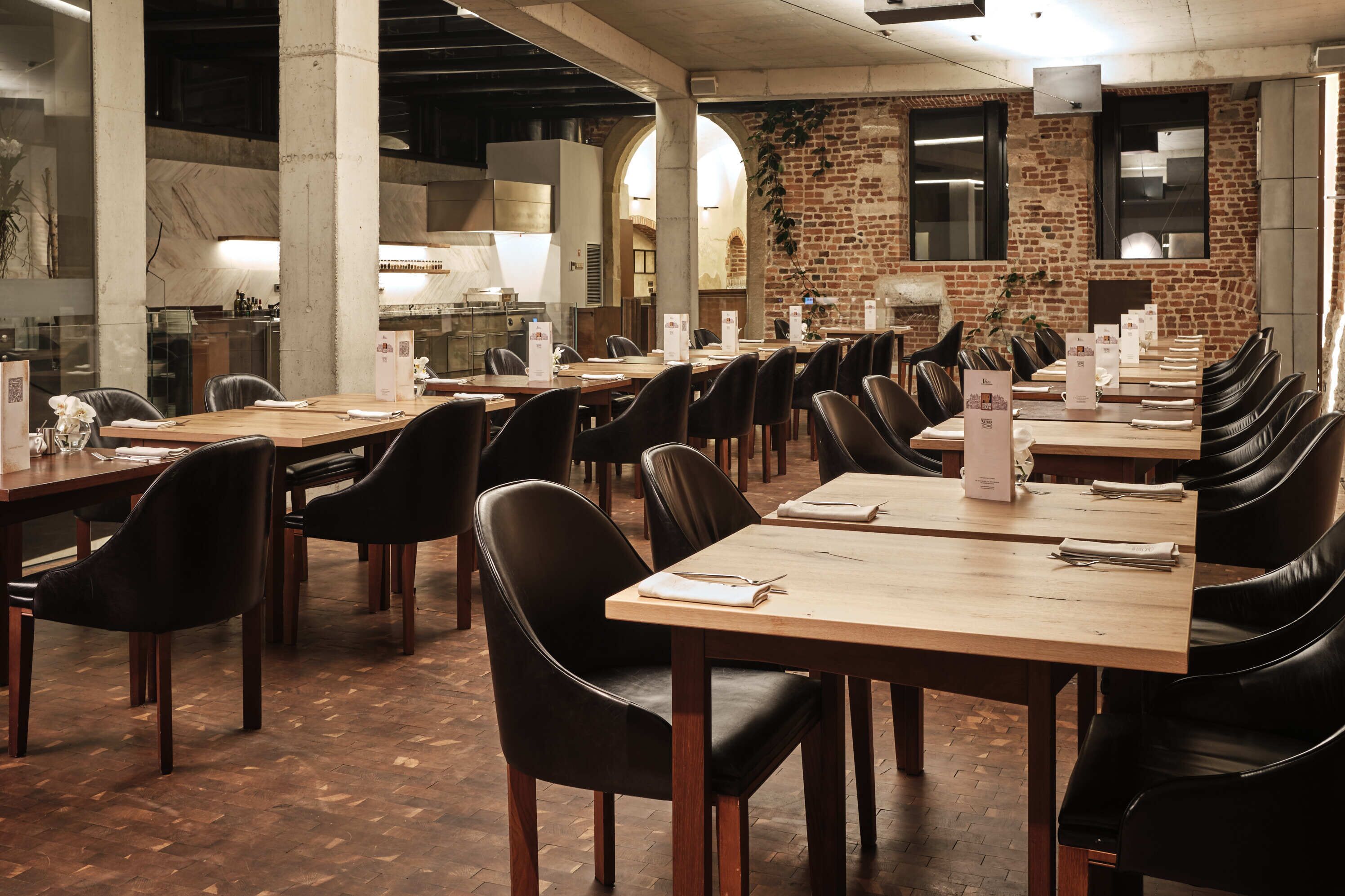 A spacious dining area with long tables and black leather chairs, featuring a brick wall and large windows.