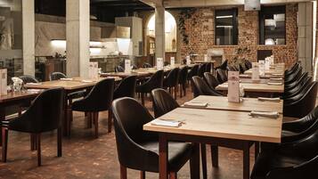 A spacious dining area with long tables and black leather chairs, featuring a brick wall and large windows.