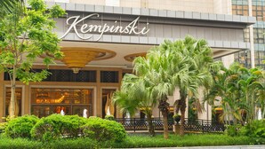 Ein Gebäude mit einem Schild, das 'Kempinski' zeigt, und einem gepflegten Garten mit Palmen.