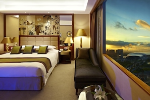 Ein Hotelzimmer mit einem großen Bett, einer Sitzecke, einem Nachttisch, einer Lampe und Blick auf eine Stadtlandschaft.