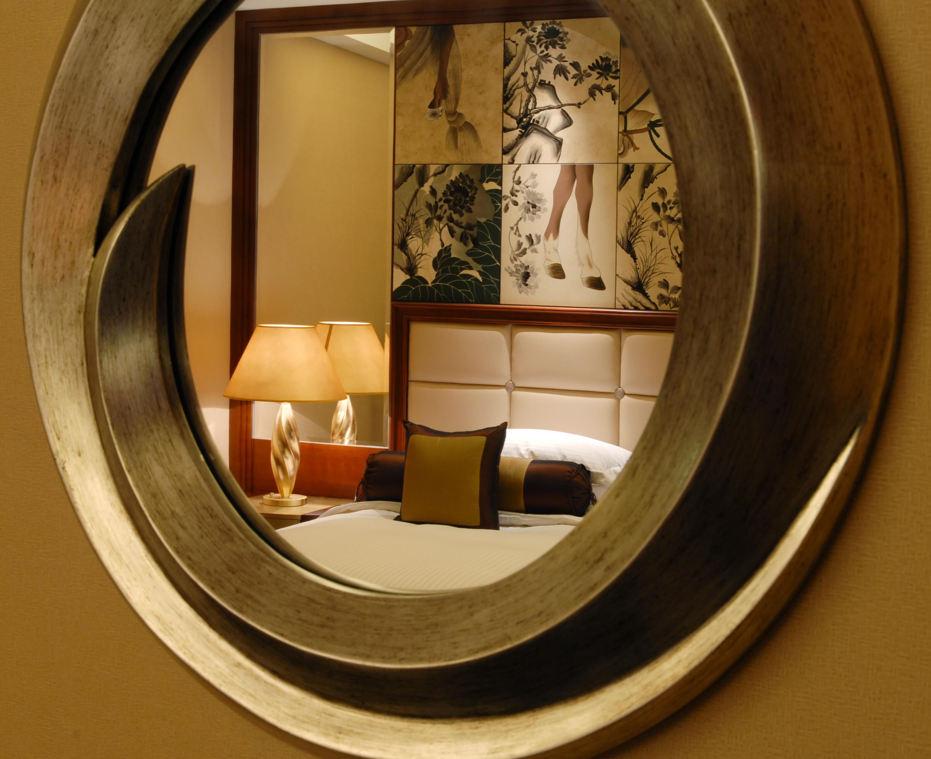 A circular mirror reflecting a room with a bed, a nightstand with a lamp, and a decorative wall panel.