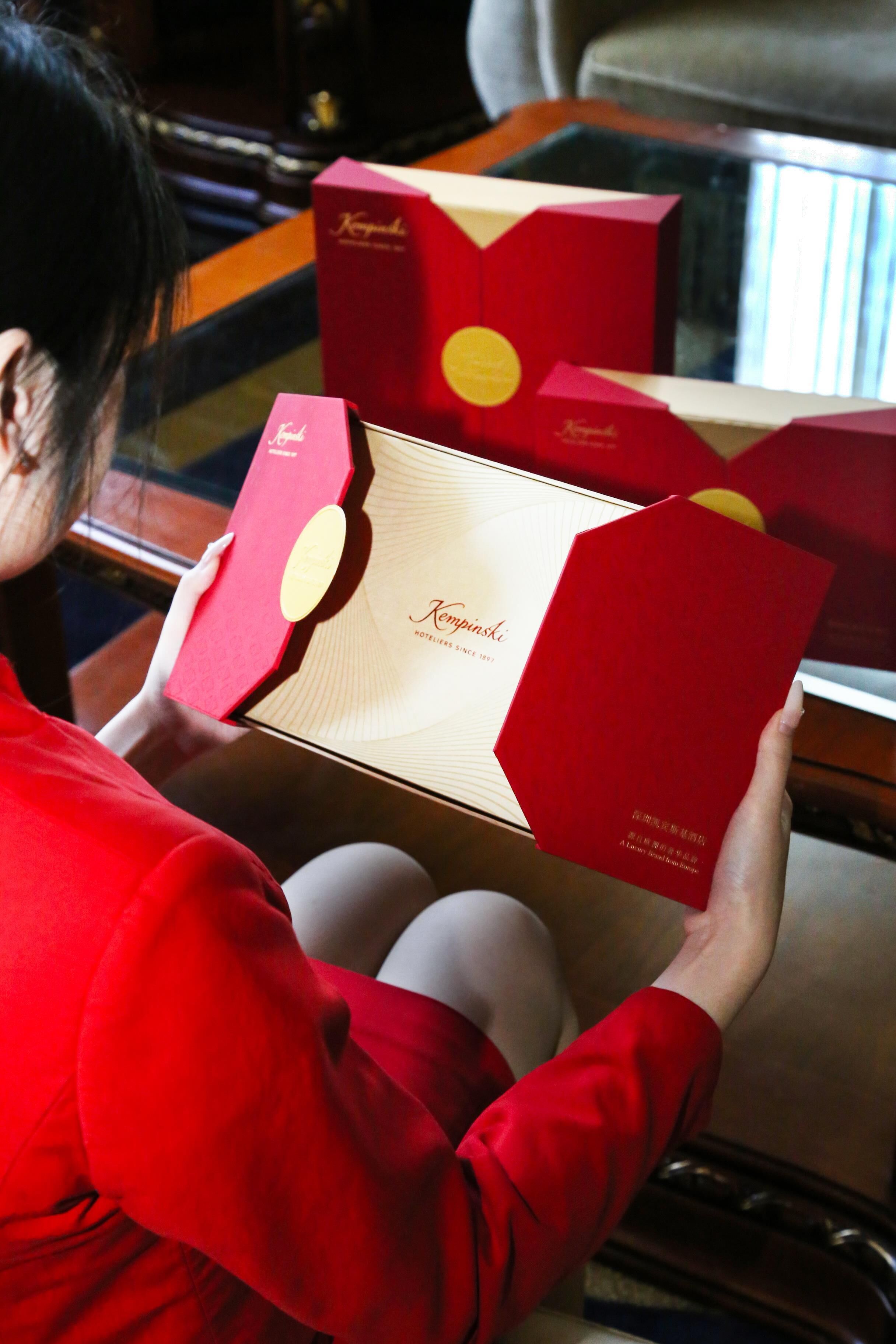 A person holding a red box with a logo, seated at a table with a reflective surface.