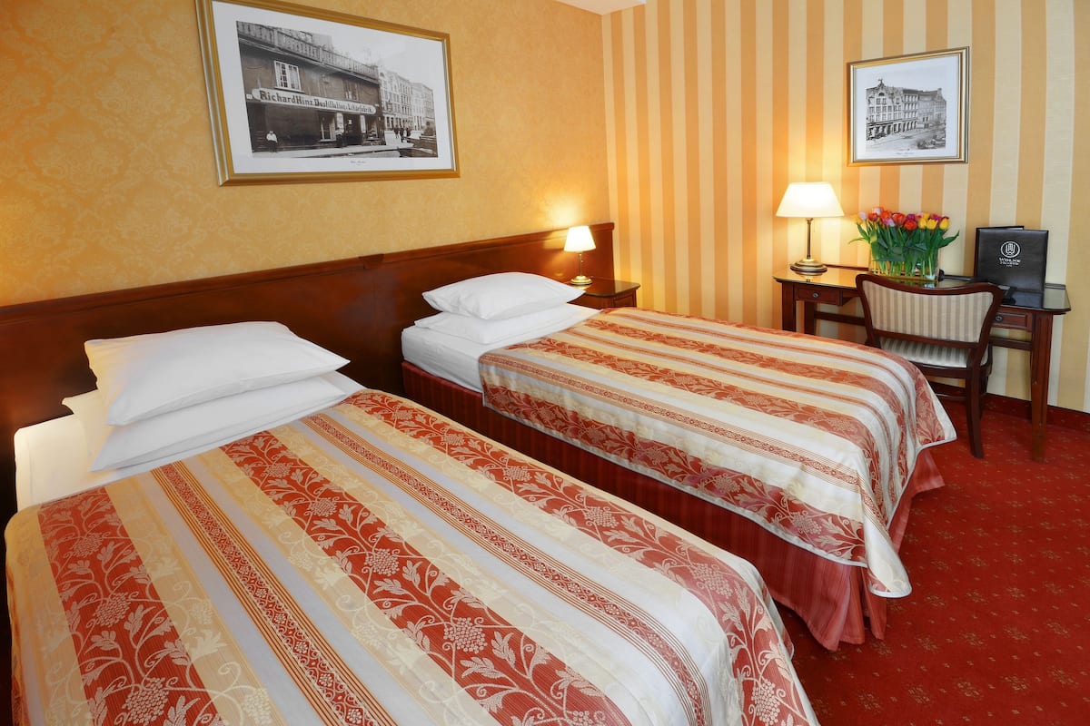 A hotel room with two beds, a desk, a chair, a lamp, and two framed pictures on the wall.