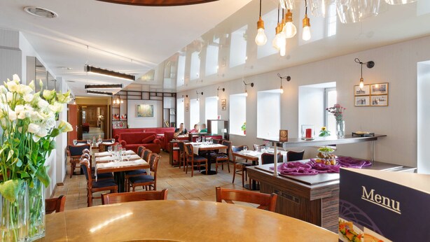 A spacious restaurant with high ceilings, wooden paneling, and modern lighting fixtures. Tables are arranged for dining, and there is a visible menu stand.
