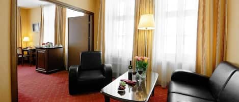 Habitación de hotel con alfombra roja, sofá de cuero negro, mesa de centro de vidrio con un florero y una lámpara, además de vistas a través de cortinas translúcidas.