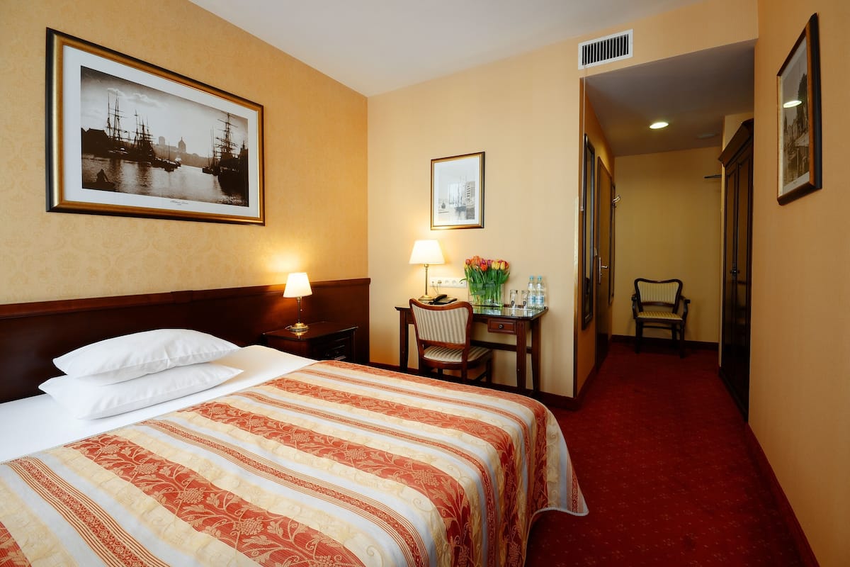 A hotel room with a large bed, a desk with a lamp, a chair, and framed pictures on the walls.
