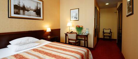 A hotel room with a large bed, a desk with a lamp, a chair, and framed pictures on the walls.