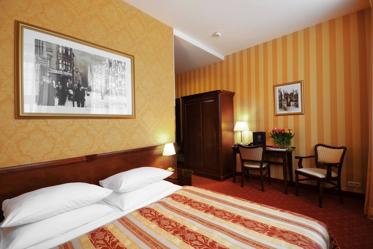 A hotel room with a large bed, a desk with a chair, a wardrobe, and two framed pictures on the wall.