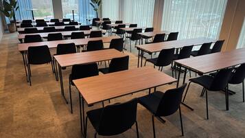 Un salón de conferencias con varias mesas y sillas, amplias ventanas con cortinas y una planta en maceta.