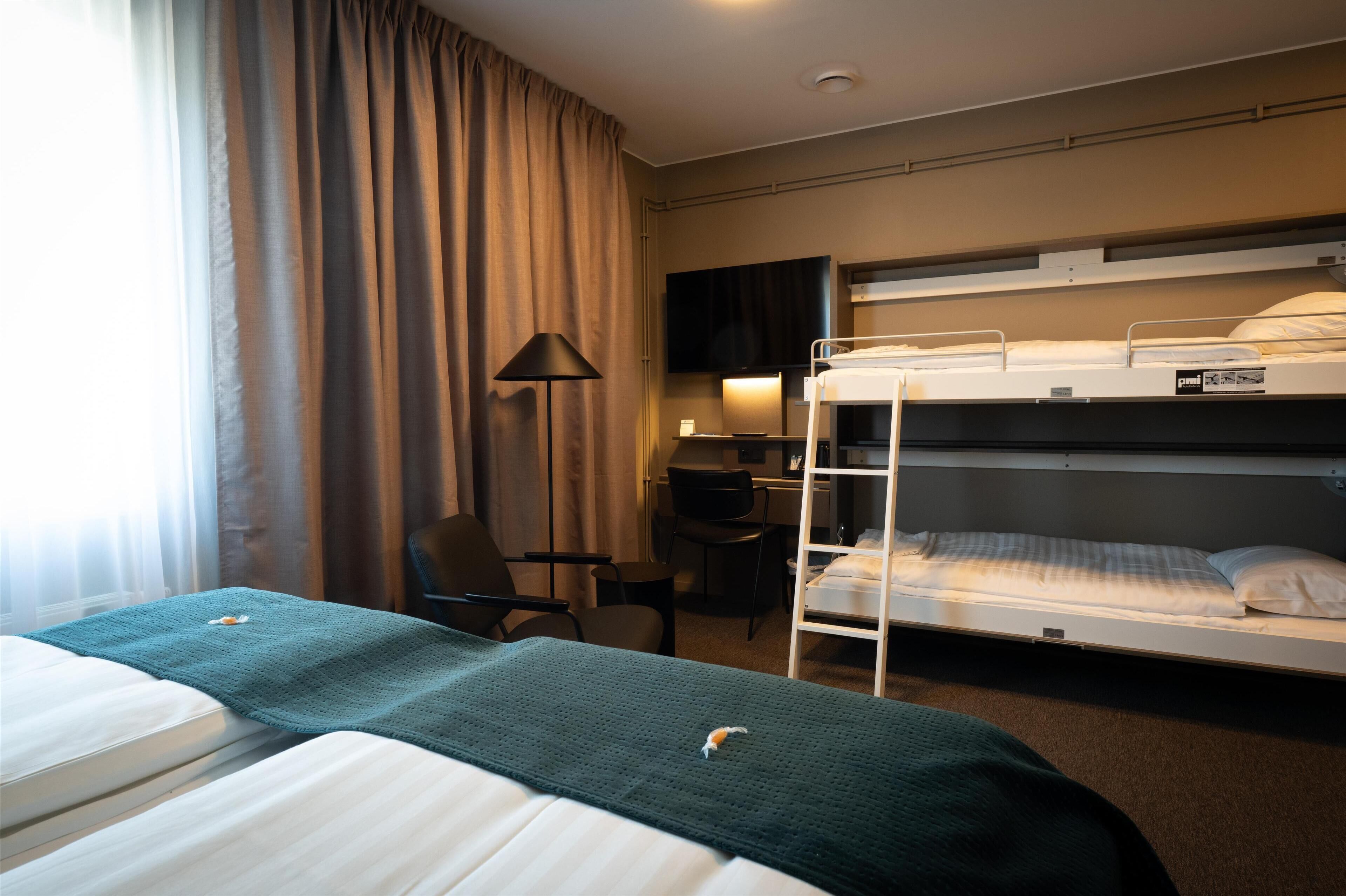Et hotelværelse med to senge, et skrivebord, en stol, et fjernsyn og et vindue med gardiner.