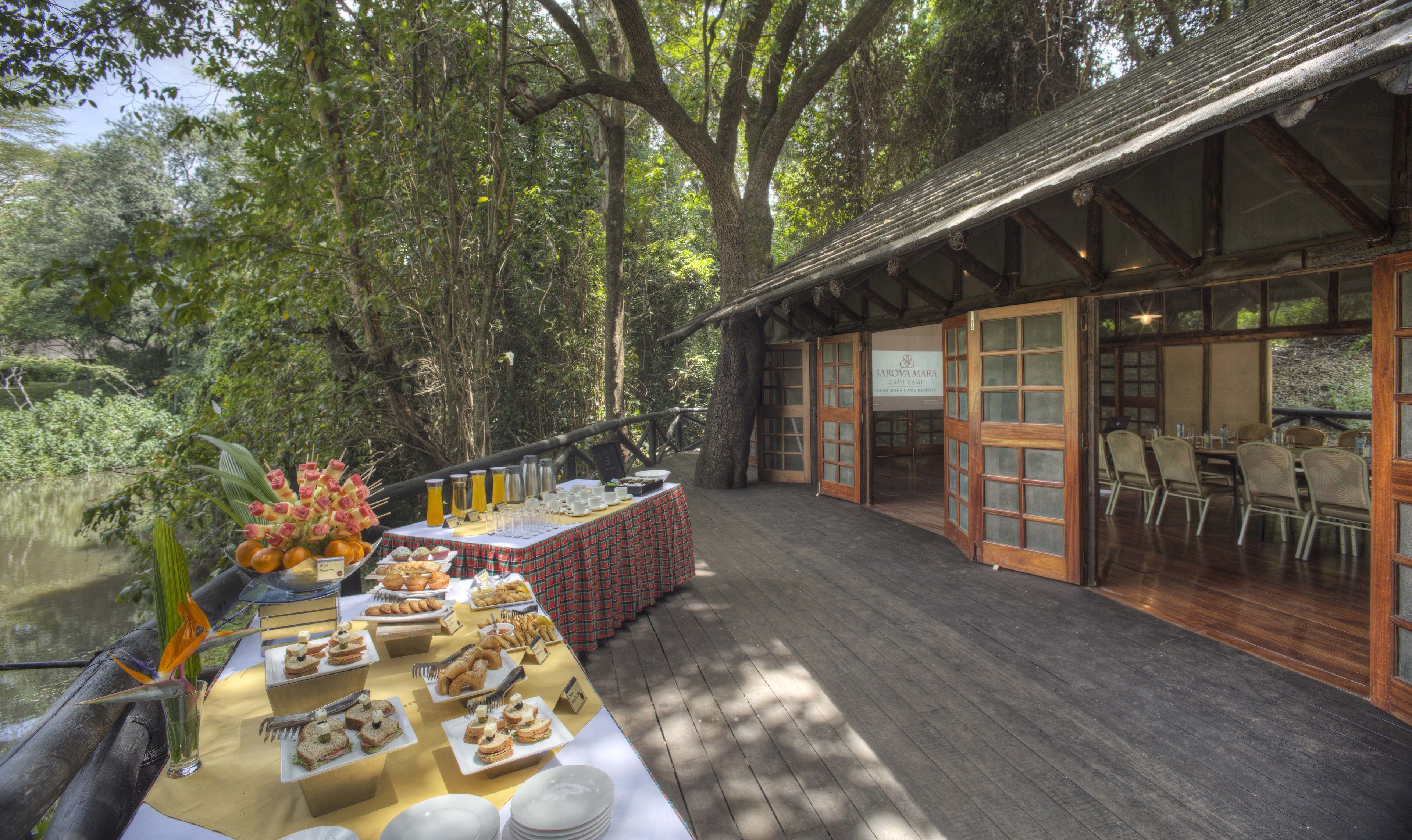 Un área de comedor al aire libre con mesas de buffet, rodeada de árboles y una terraza de madera.