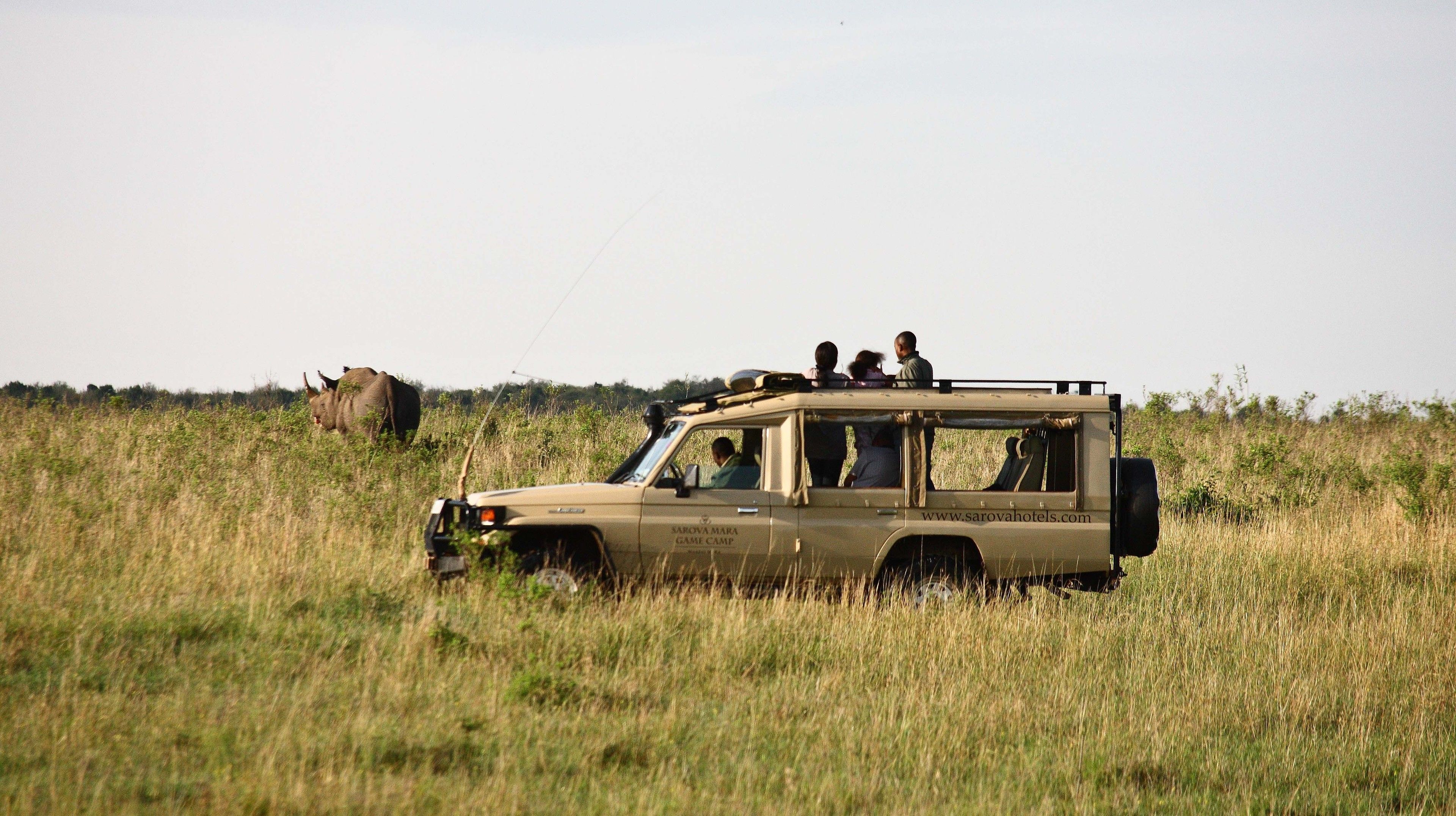 Un véhicule de safari transportant des passagers, garé dans un champ herbeux, avec un grand rhinocéros en arrière-plan.