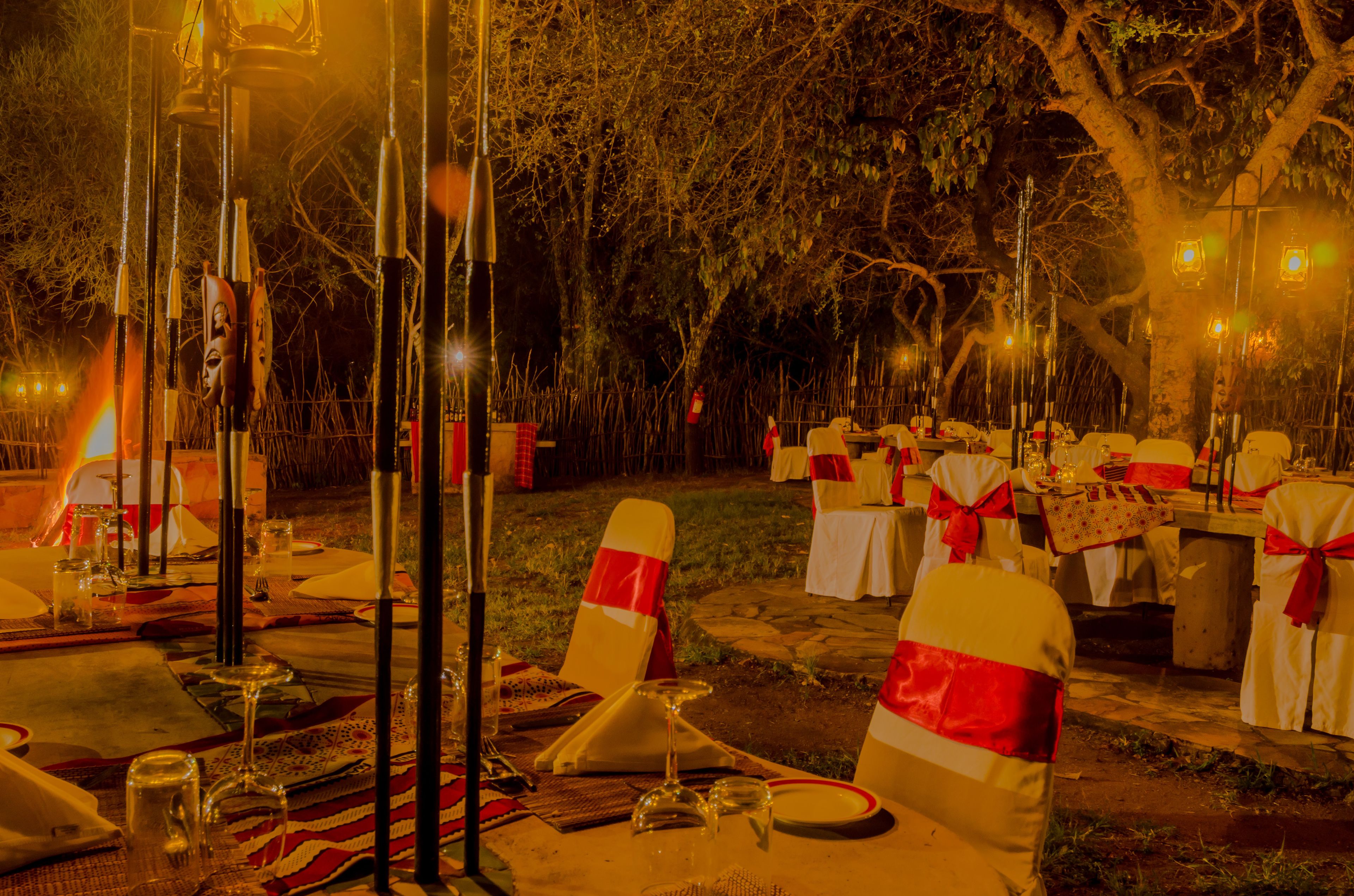 An outdoor dining area with tables set for a meal, chairs with red sashes, and a warm ambient light.
