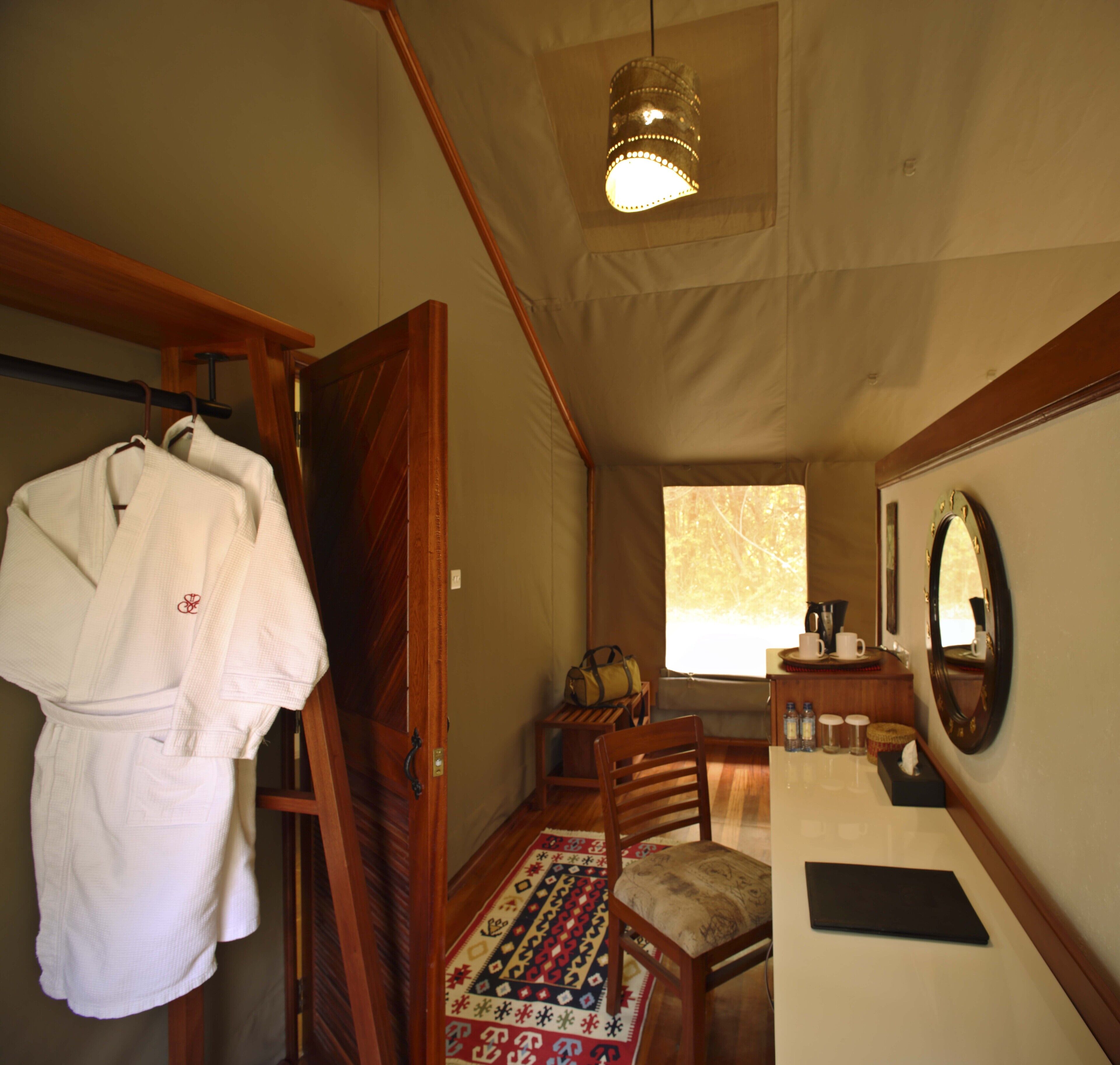 A room with a wooden door, a white robe hanging, a patterned rug, a wooden chair, a small table with a mirror, and a ceiling light.