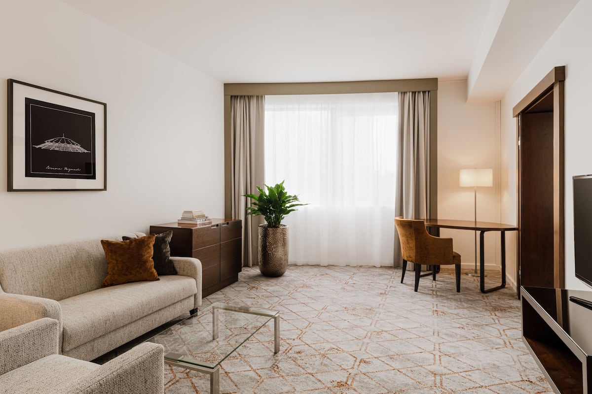 Ein modernes Hotelzimmer mit Sofa, Couchtisch, Fernseher, Esstisch mit Stühlen, einer Zimmerpflanze und einem großen Fenster mit Vorhängen.
