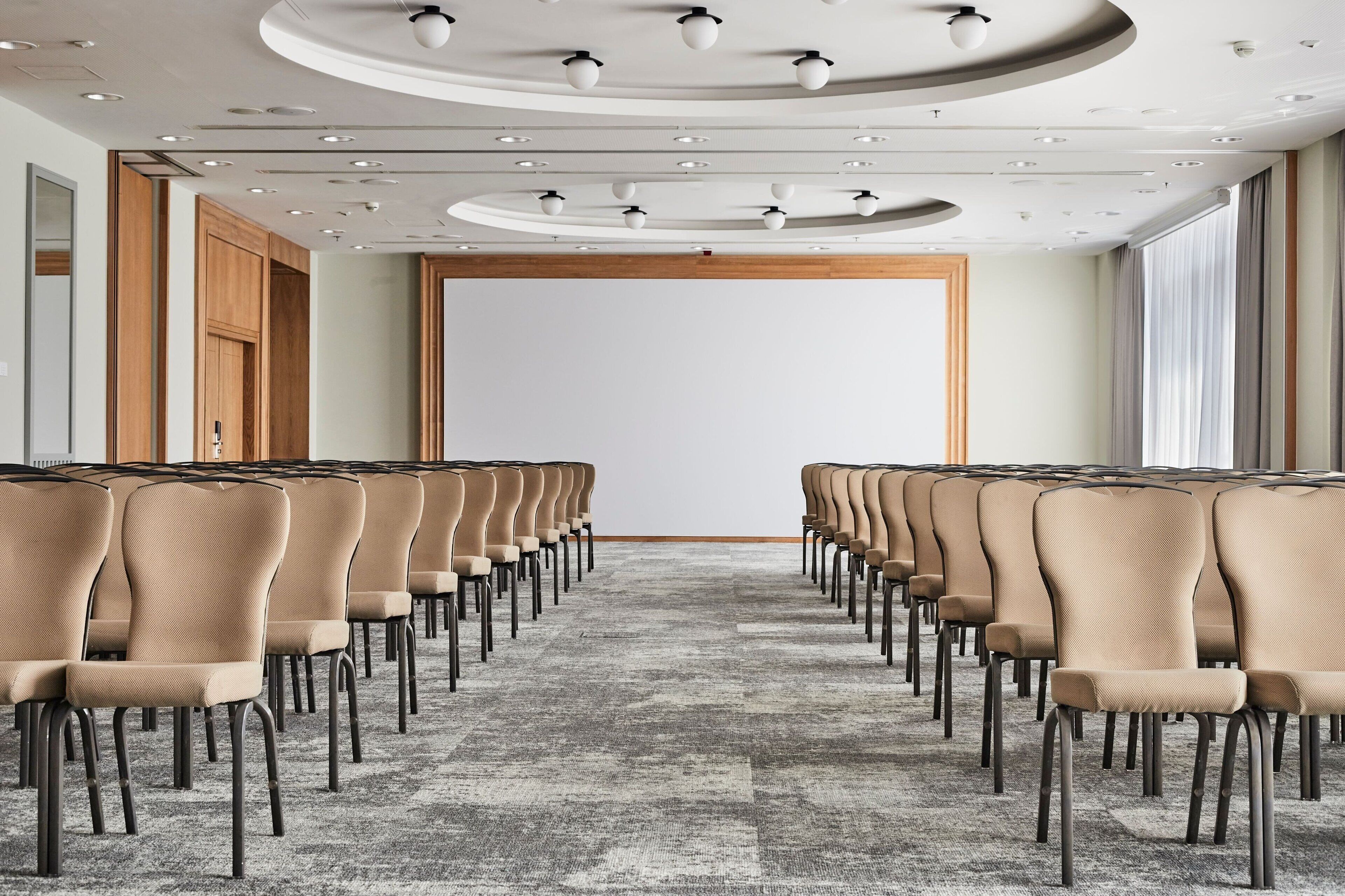 Un salón de conferencias con filas de sillas color beige, suelo alfombrado y una pantalla blanca grande al frente.