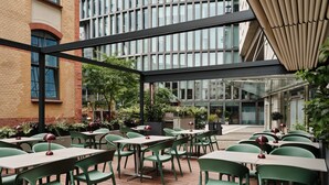 Ein Außenbereich eines Cafés mit grünen Stühlen und Tischen, umgeben von Ziegelgebäuden und viel Grün.