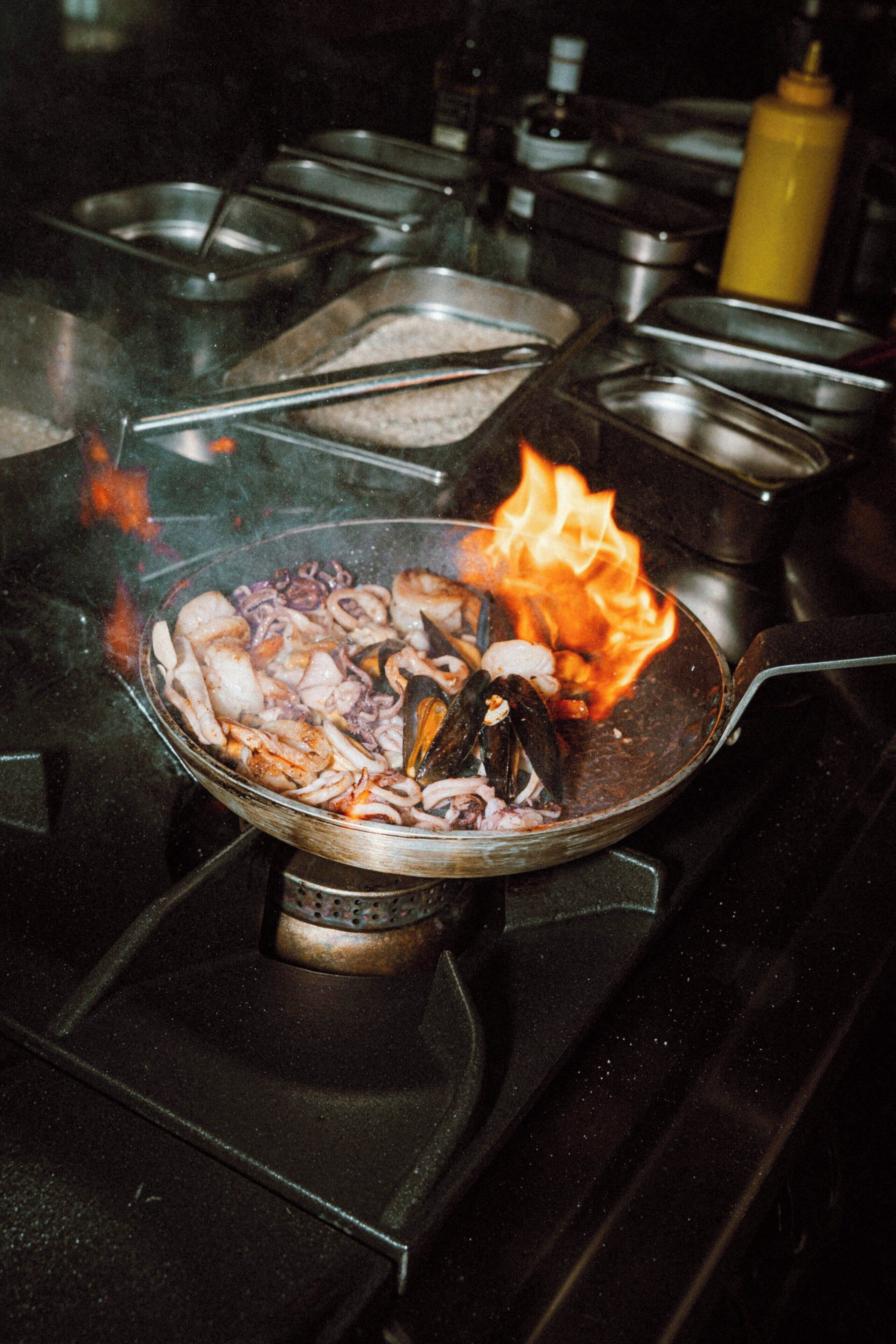 A kitchen with a pan on a stove, containing seafood and flames.
