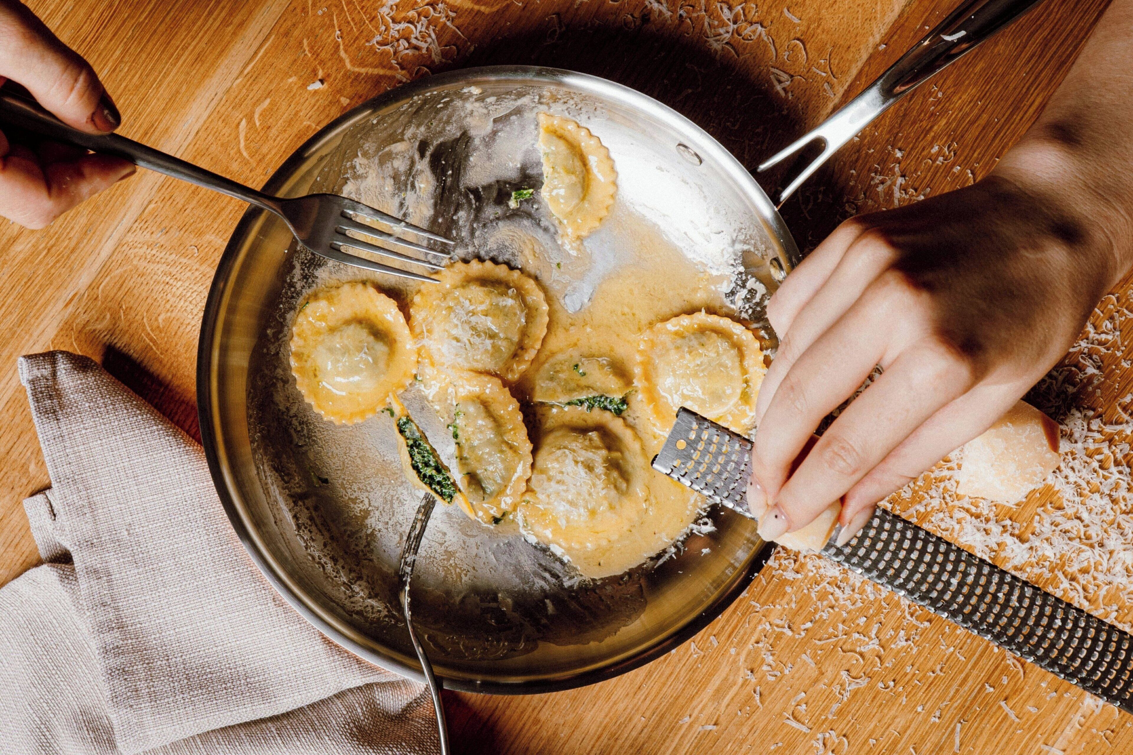 Eine Person reibt Käse an einer Reibe, in der Nähe steht ein Topf mit Ravioli.
