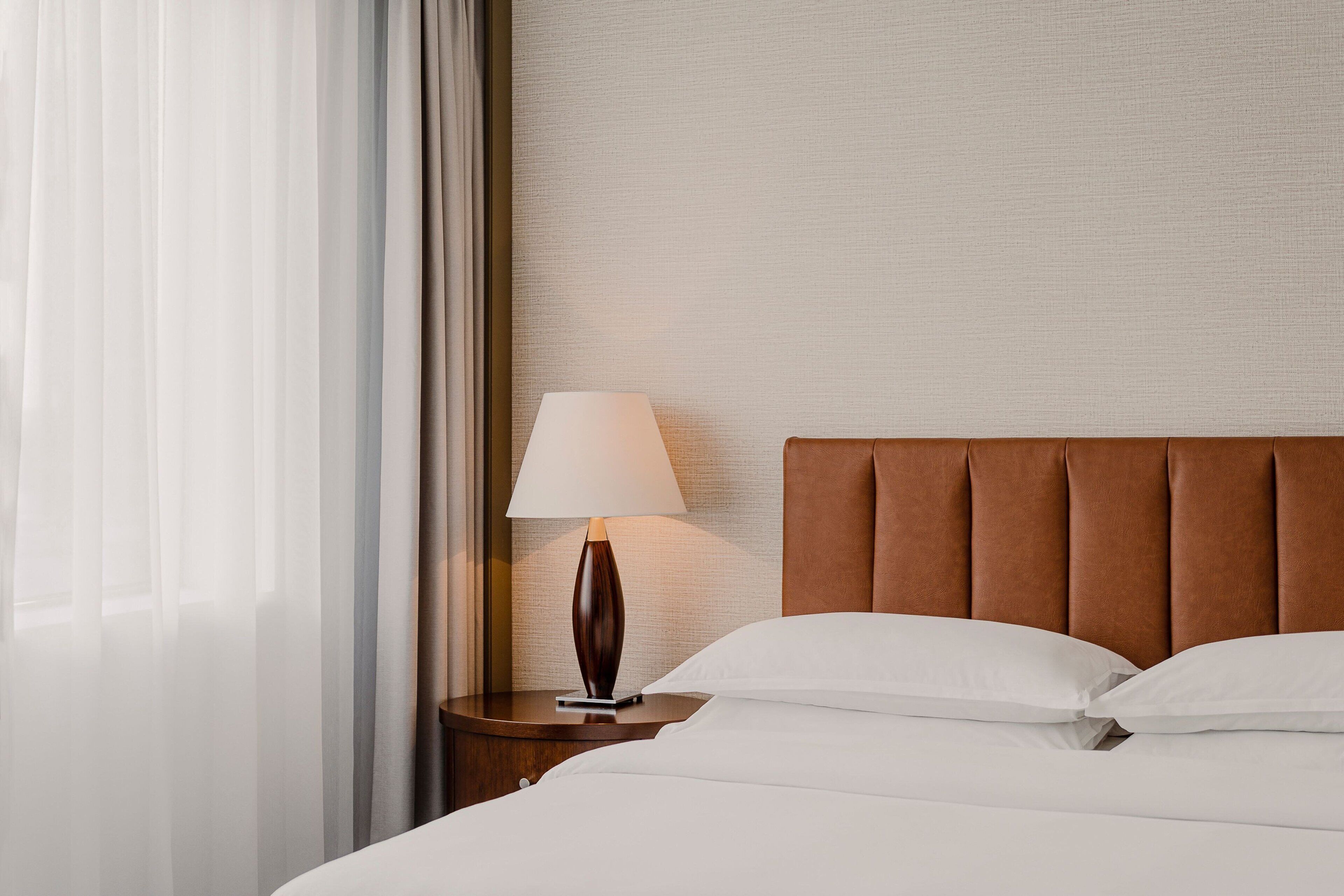 A neatly made bed with white linens, a brown upholstered headboard, and a bedside lamp on a wooden nightstand.