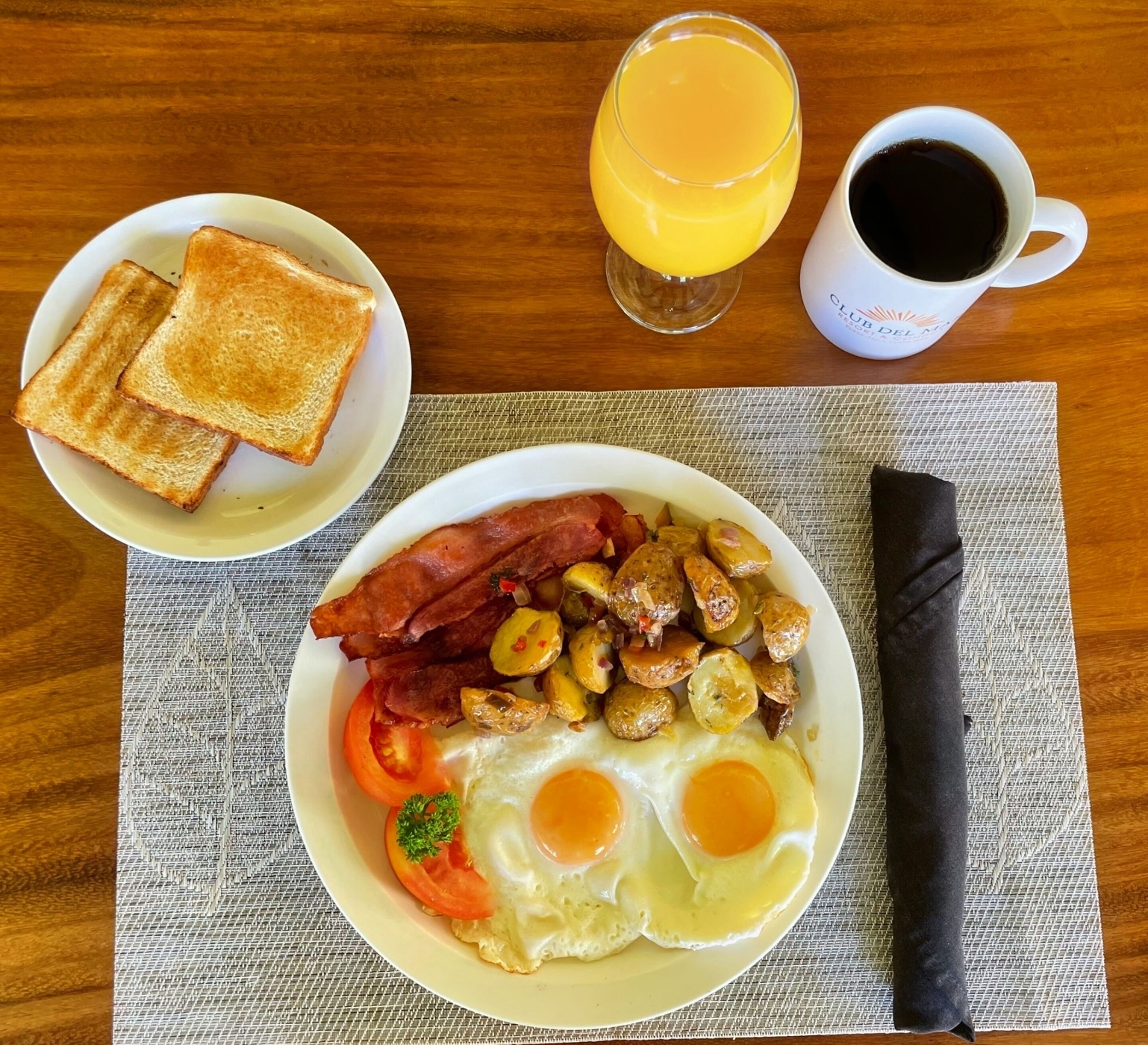 Prato de café da manhã com ovos, bacon, batatas e torradas, acompanhado de um copo de suco de laranja e uma xícara de café.