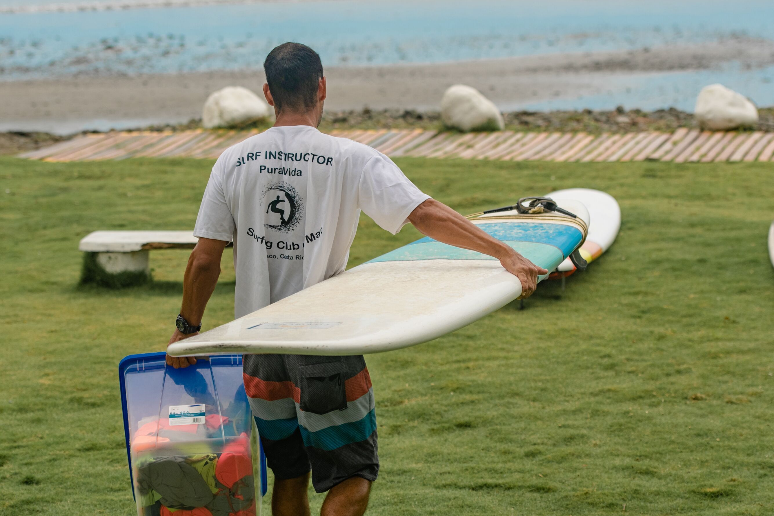 A person carrying a surfboard and a bag on a grassy area near the beach.