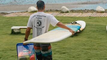 A person carrying a surfboard and a bag on a grassy area near the beach.
