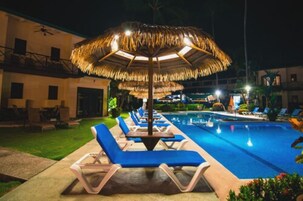 Un área de piscina con sillones de descanso y sombrillas, rodeada de edificios y vegetación.