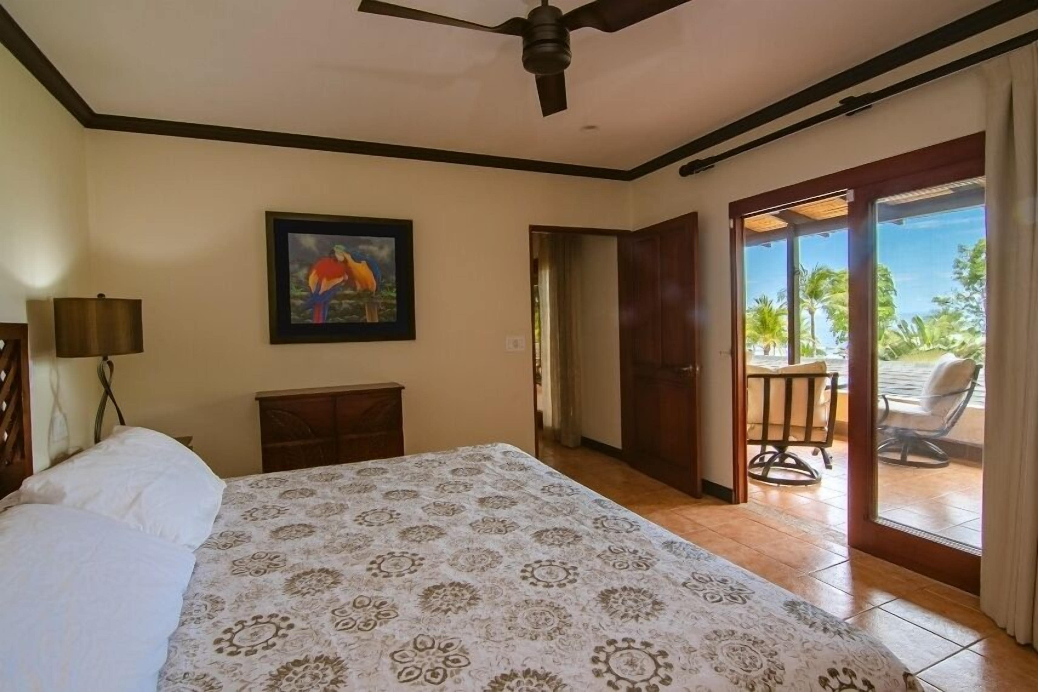 A hotel room with a bed, a ceiling fan, a painting, and a view of the outdoors through a sliding door.