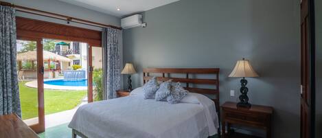 A bedroom with a large bed, two bedside tables with lamps, a wooden headboard, and a view of a swimming pool and outdoor area.