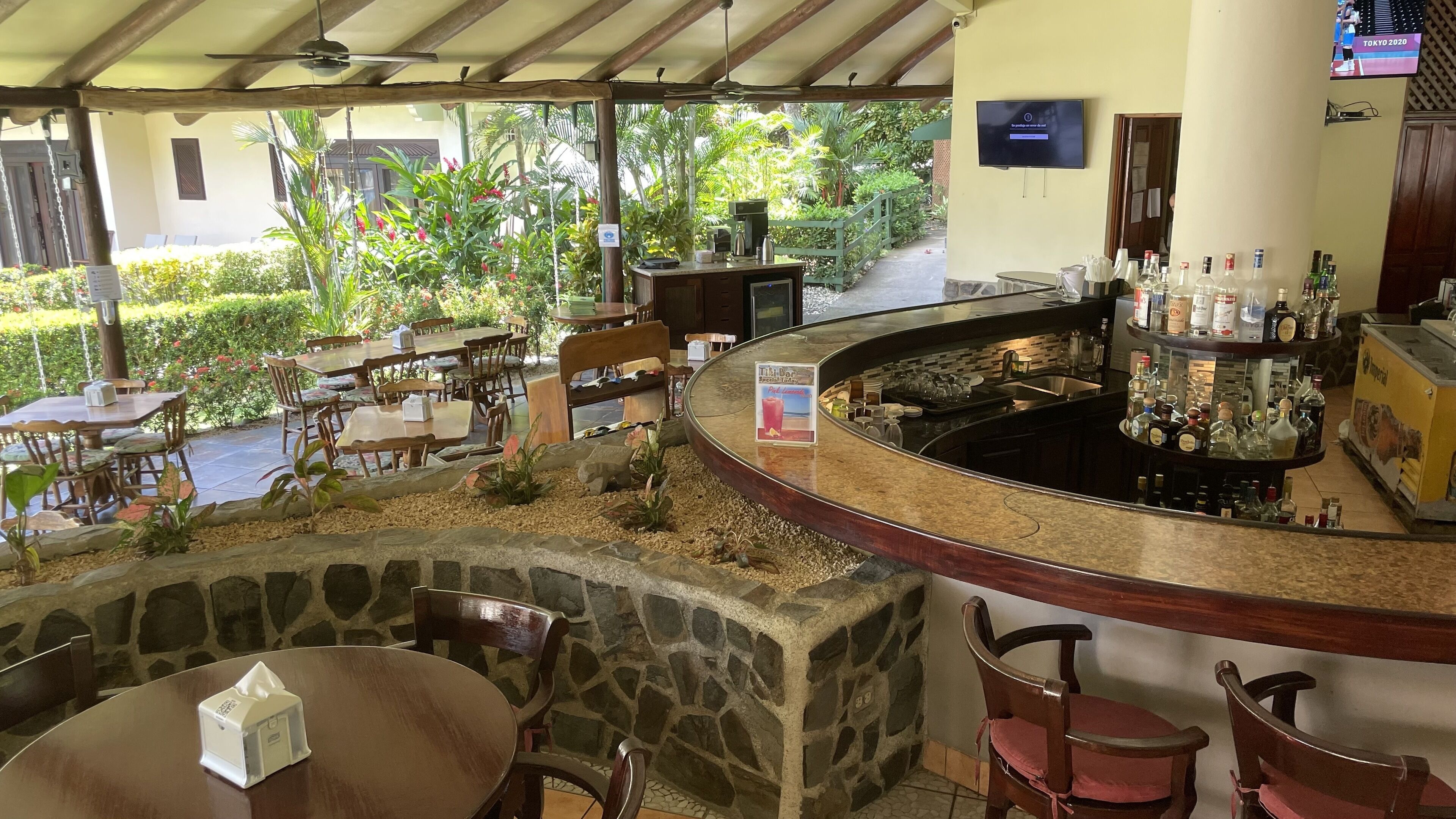 Zona de bar con mostrador de piedra, sillas de madera y vista a un jardín con plantas variadas y un sendero.