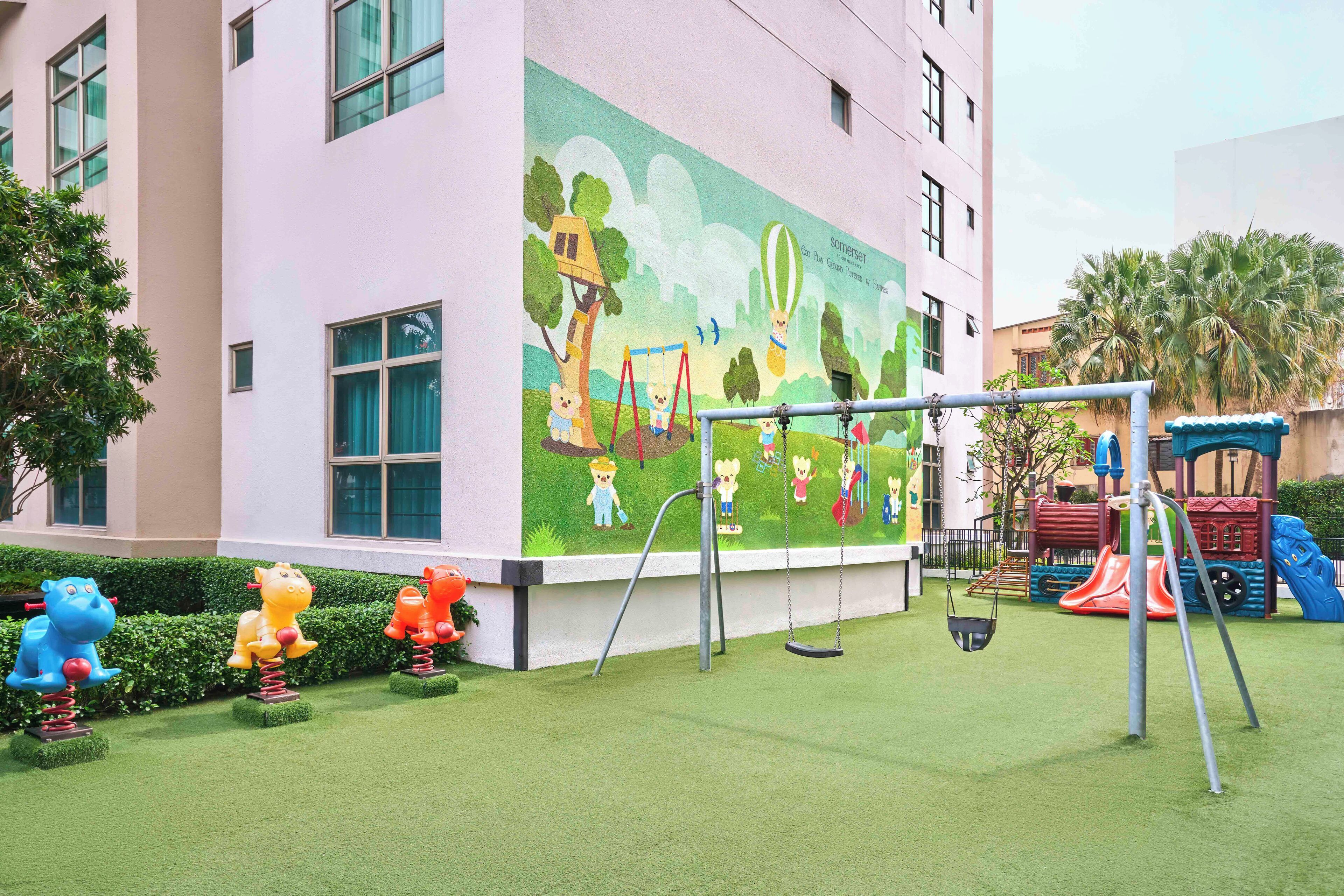 A children's playground with swings and a slide, adjacent to a building with a mural of a park scene.