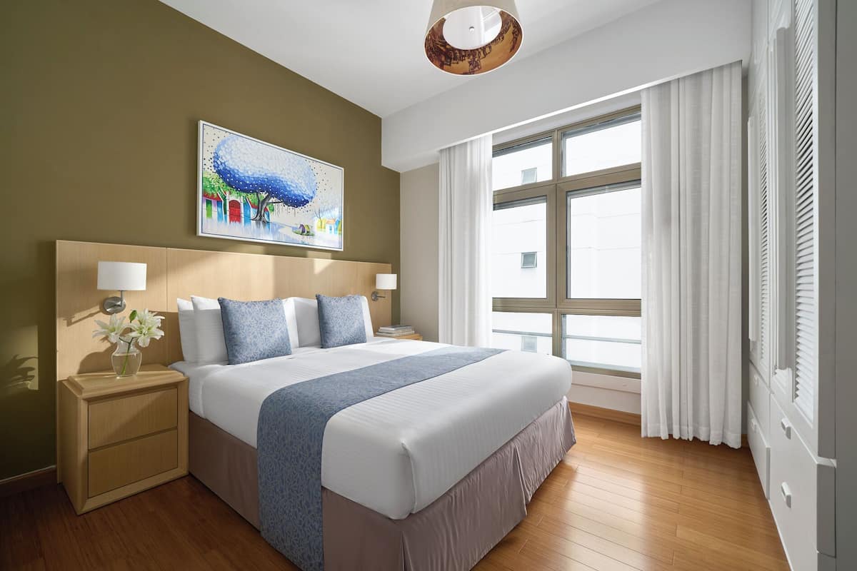 Una habitación de hotel moderna con una cama grande, dos mesitas de noche con lámparas, un cuadro en la pared y un ventanal con cortinas.