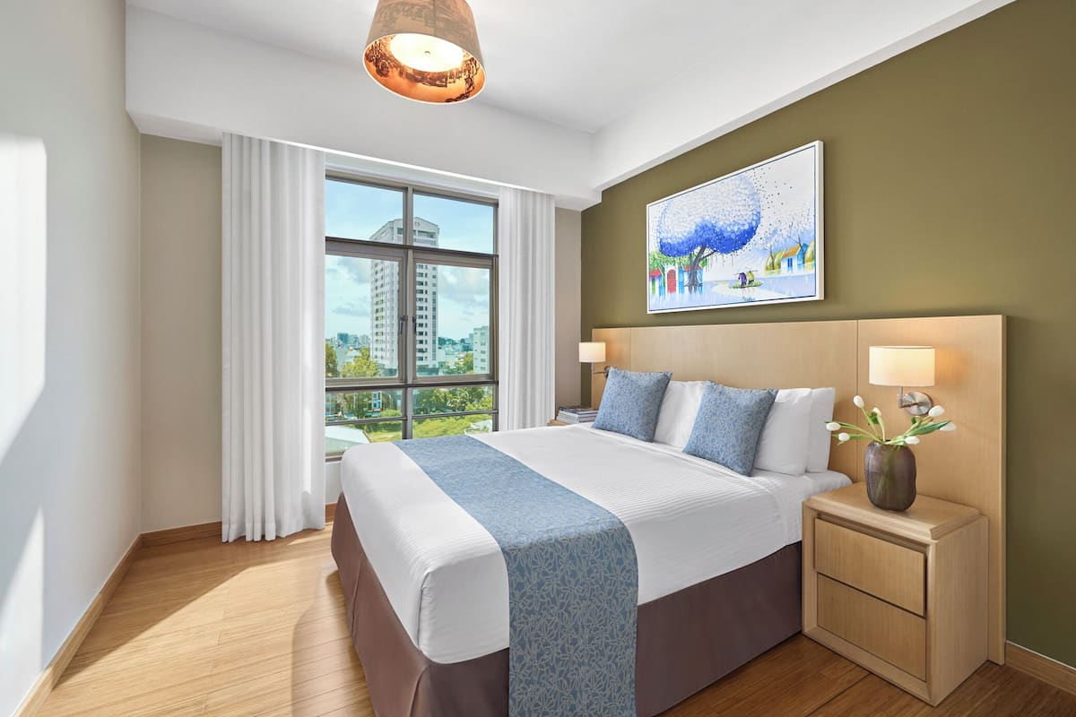 Una habitación de hotel moderna con una cama grande, una mesita de noche con lámpara, un cuadro en la pared y una ventana con cortinas.