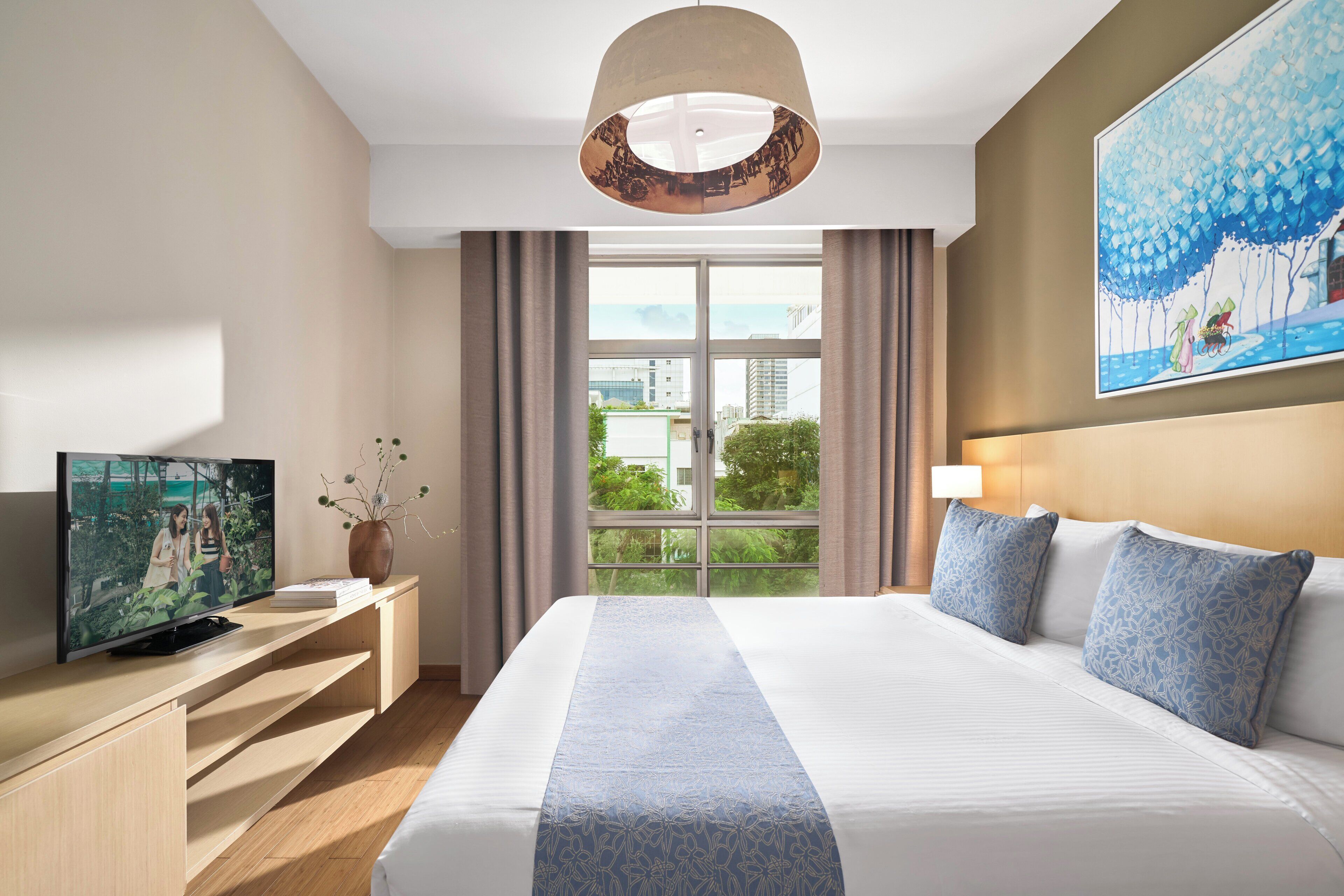 A hotel room with a large bed, a flat-screen TV on a wooden console, a vase with flowers, a painting on the wall, and a view of greenery through the window.