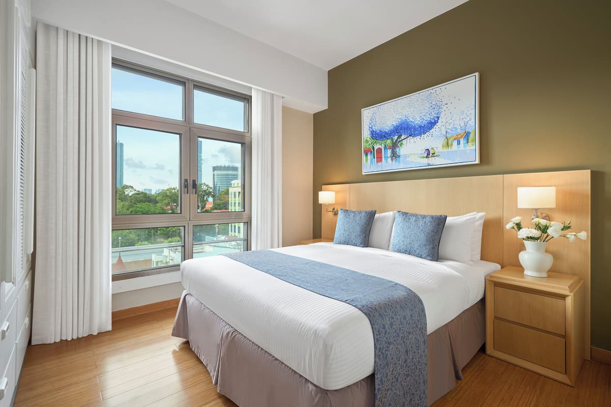 Habitación de hotel con una cama grande, una mesita de noche con un florero, un cuadro en la pared y vistas a la ciudad a través de la ventana.