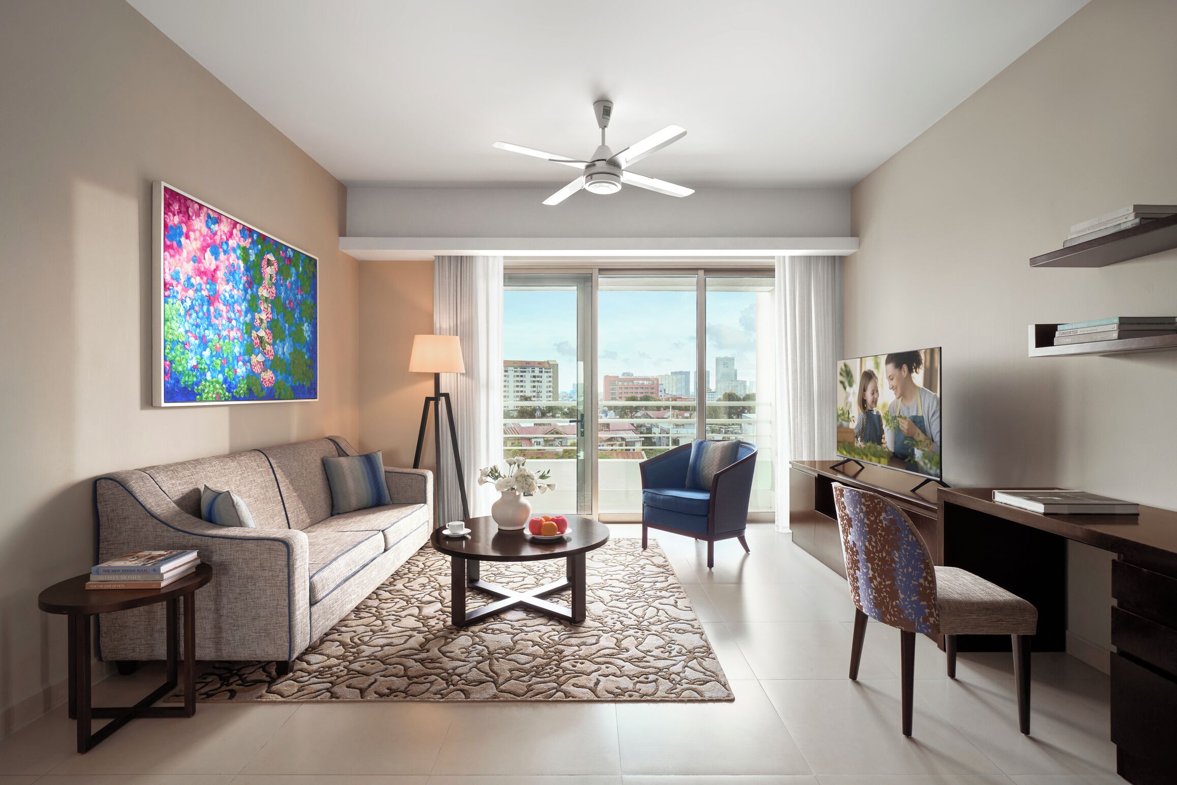 A modern living room with a sofa, coffee table, and chairs, a ceiling fan, and a large window with a city view.