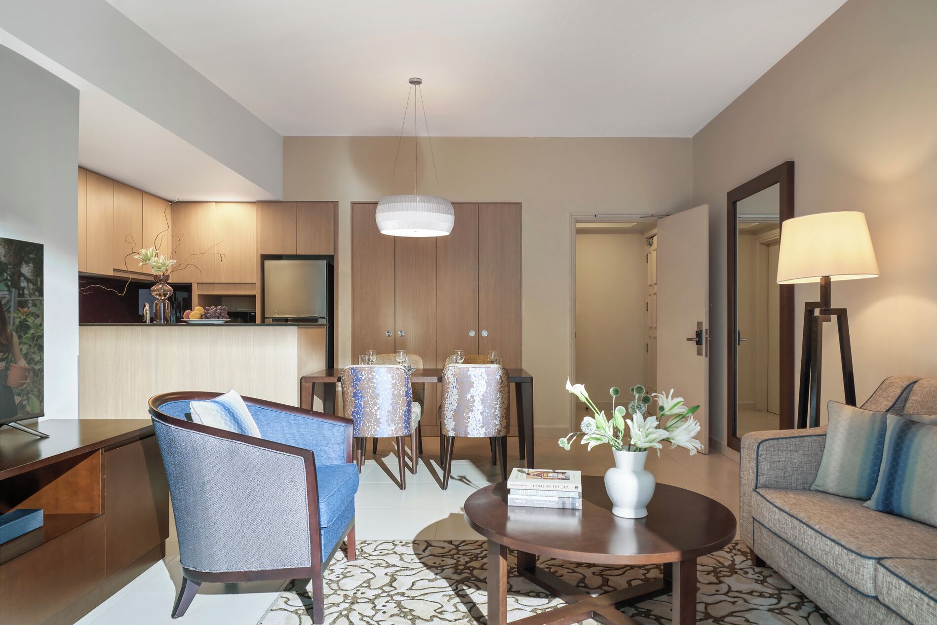 A modern living room with a sofa, two chairs, a coffee table, and a dining area with a table and chairs. There is a kitchen area with a refrigerator and cabinets, and a large mirror on the wall.