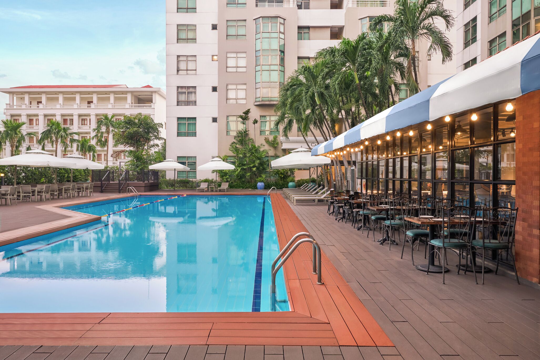 A swimming pool with a wooden deck, surrounded by lounge chairs and umbrellas, with a restaurant area featuring outdoor seating and a view of a tall building.