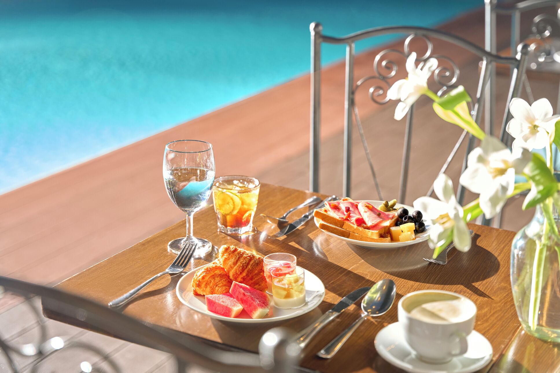 A breakfast setting by a poolside with a plate of pastries, a glass of water, a fruit drink, and a cup of coffee.