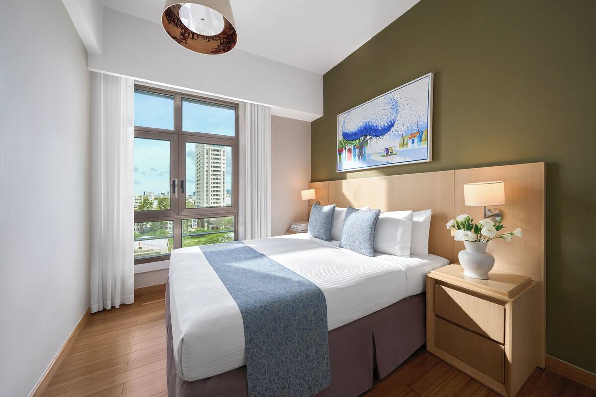 Una habitación de hotel moderna con una cama grande, una mesita de noche con un florero y un cuadro en la pared, además de una ventana con cortinas.