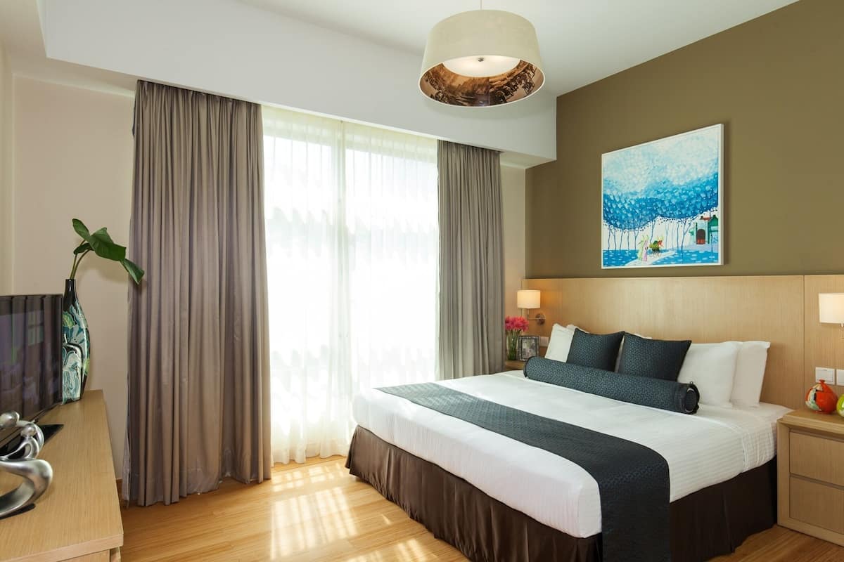 Una habitación de hotel con cama, cortinas, un cuadro, una lámpara, una mesita de noche y un televisor.