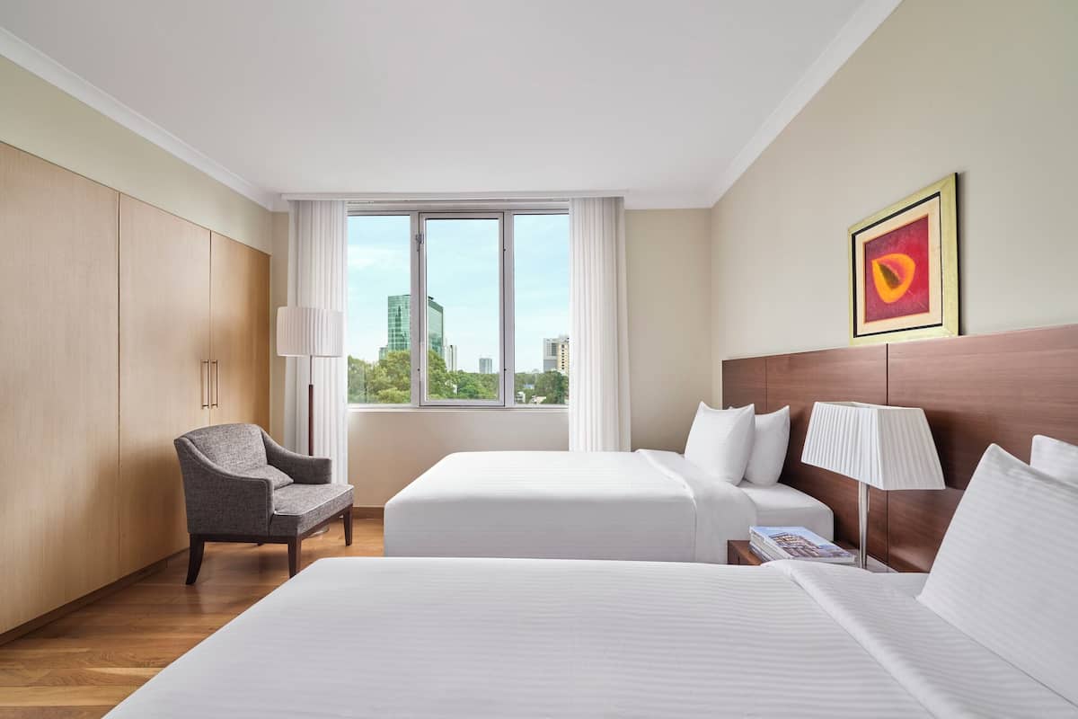 A hotel room with two beds, a chair, a window with a city view, and a framed picture on the wall.